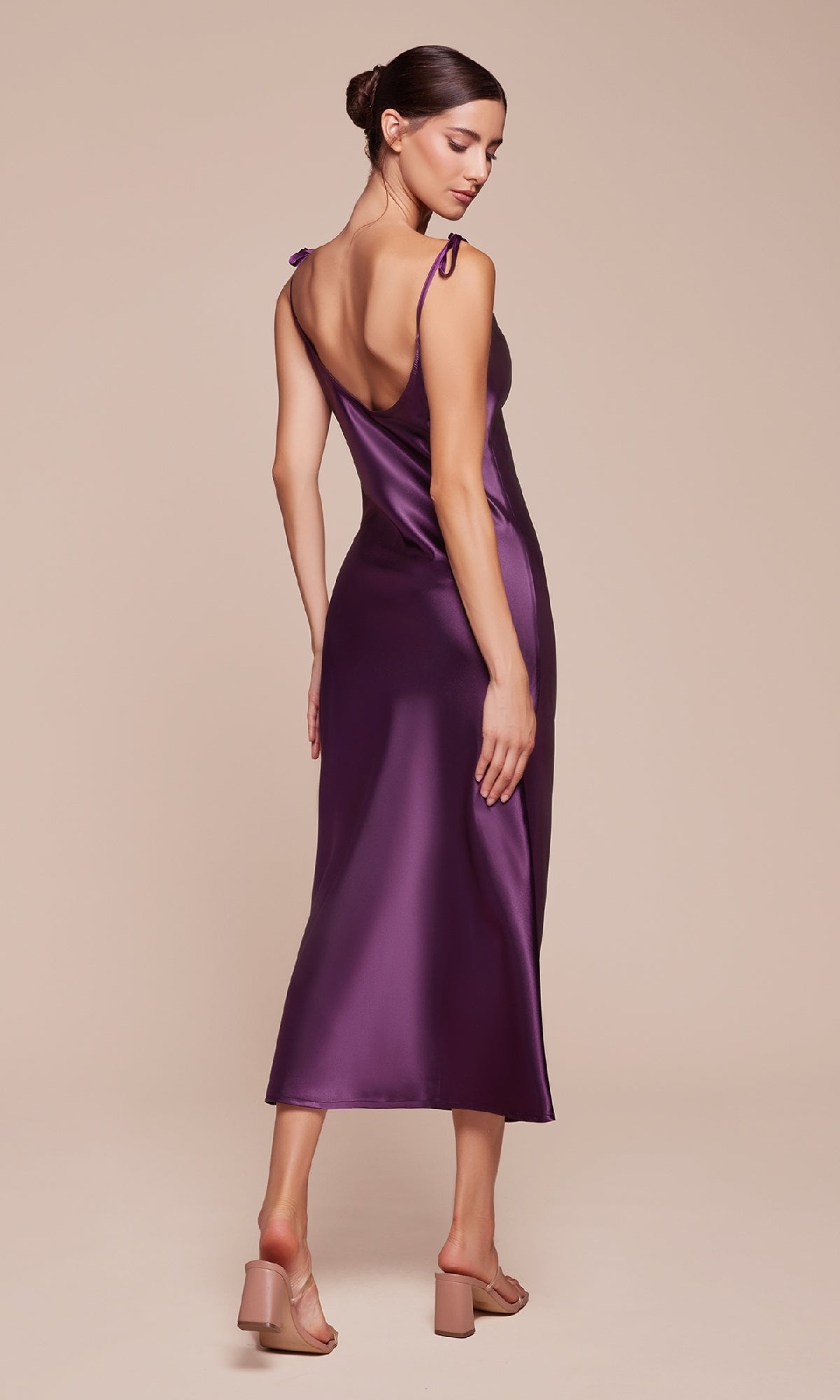 A woman in the Midi Wedding-Guest Slip Dress BD103, a sleeveless deep purple midi with tie straps, pairs it with nude block-heeled sandals as she stands facing away and looks over her shoulder.